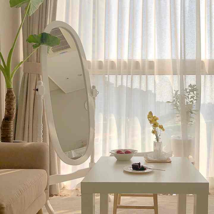 柔らかなライトベージュ色調のワンルーム、カーテン横にソファとテーブル、姿見と植物が配された落ち着いたおうちカフェの雰囲気