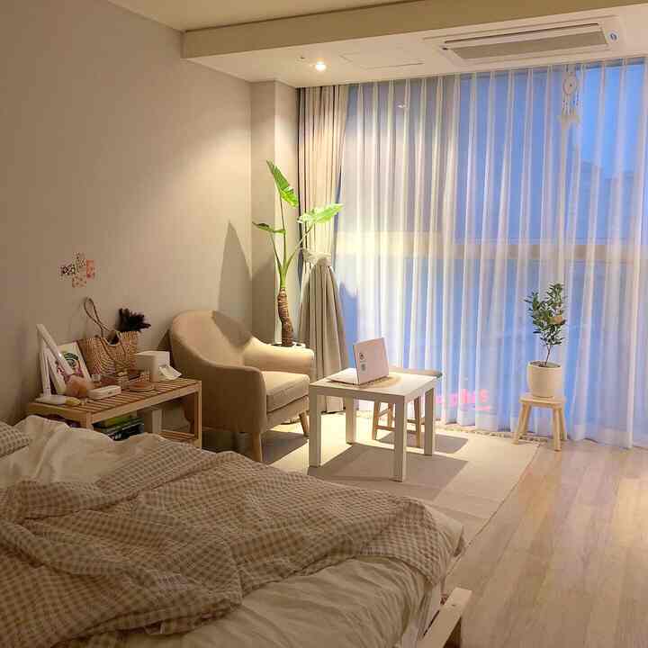 Natural toned bedroom featuring a beige armchair, white table, curtains, and plants creating cozy atmosphere
