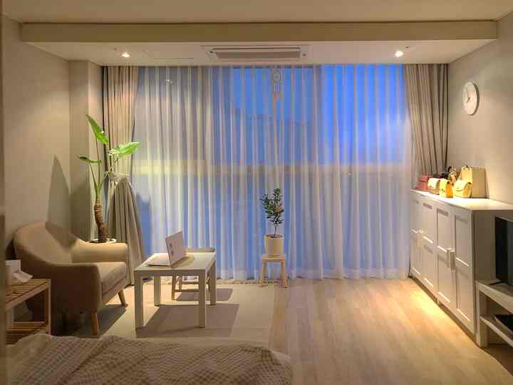Cozy studio living room in bright white and beige tones featuring a white coffee table with wooden stool, beige armchair and plants on left side