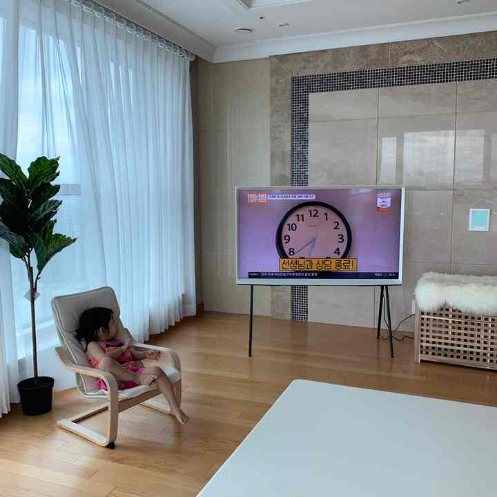 Living room with white curtains and wood tone flooring, featuring a child seated on an armchair watching TV in a natural setting
