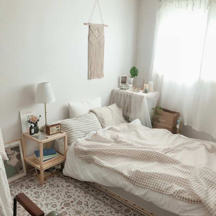 White and beige bedroom featuring natural wood side table and macrame wall hanging under soft sunlight creating a cozy atmosphere