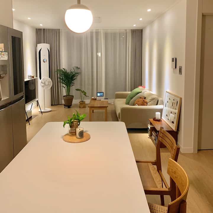 Natural-toned corridor apartment kitchen and living room featuring a large white dining table, wood-tone furniture, plants in a cozy two-person newlywed home