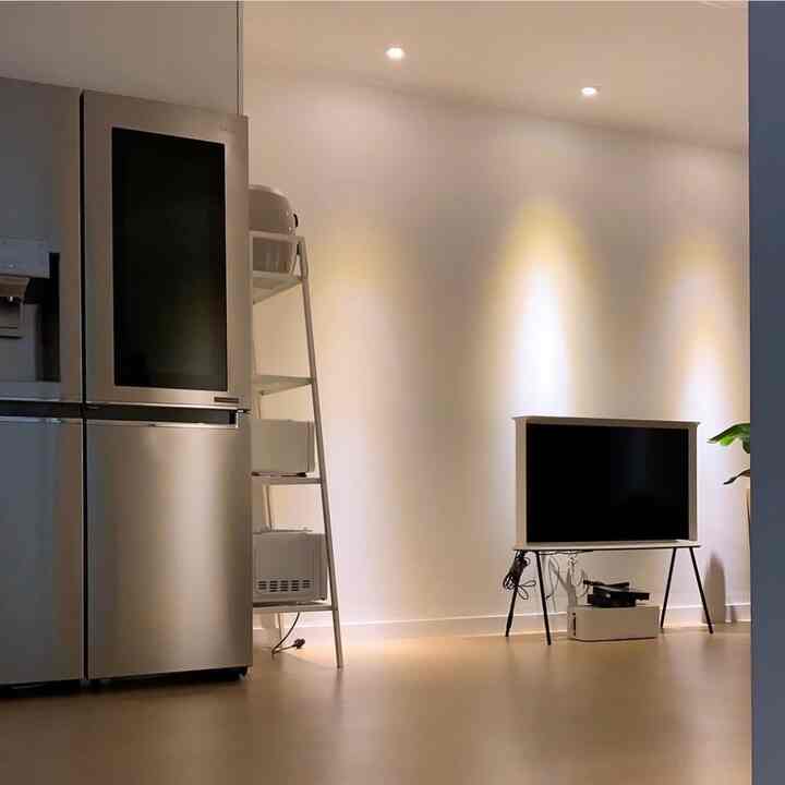 Modern living and kitchen space featuring white walls, natural wood flooring, stainless steel fridge, metal shelving, and a TV with minimalist styling