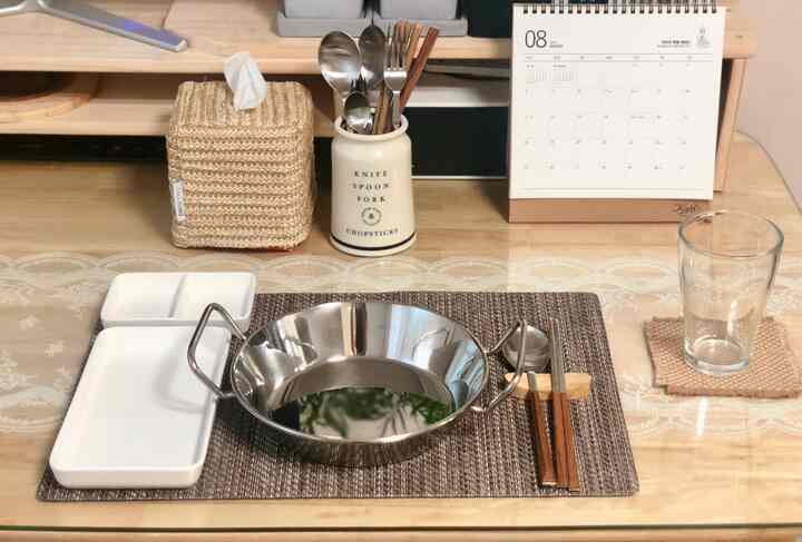 Natural wood-tone studio apartment kitchen dining table featuring ivory table runner, rattan tissue cover, and stainless steel plate set for single dining