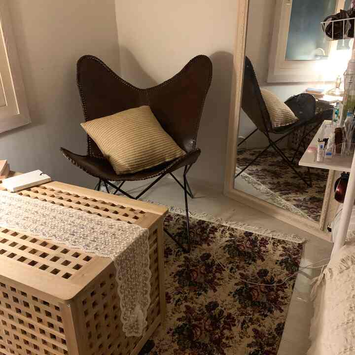 温かみのあるナチュラルトーンのワンルーム、ヴィンテージ風の花柄ラグと大型の姿見、バタフライアームチェアが配置された雰囲気のある休憩スペース