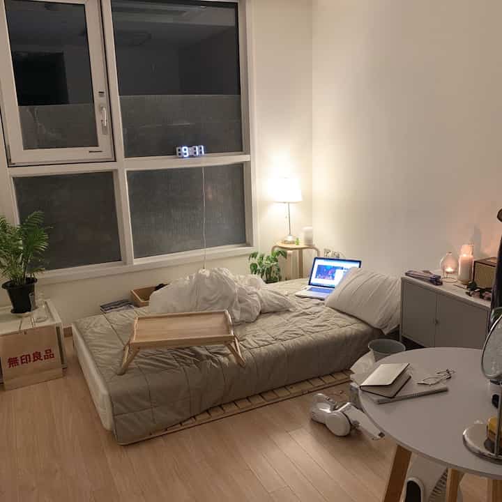 ベージュと木目調のワンルーム寝室、マットレスとトレイ、ノートパソコンとフロアランプが配置された温かみのあるインテリア