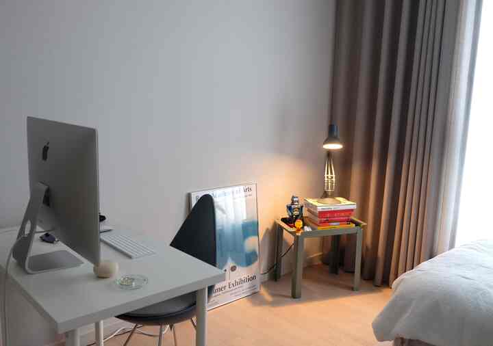 White and beige toned bedroom featuring a desk with an iMac, armchair, and table lamp creating a modern and neat home office setting