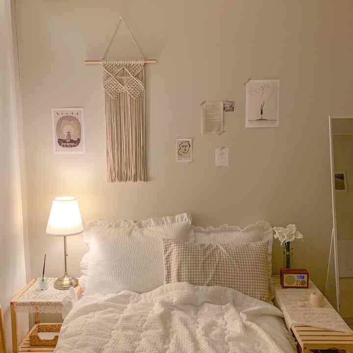 ベージュとアイボリーの色調の寝室、ベッドとナイトテーブル、壁に掛けられたマクラメとポスターが温かみのある雰囲気を作り出すインテリア