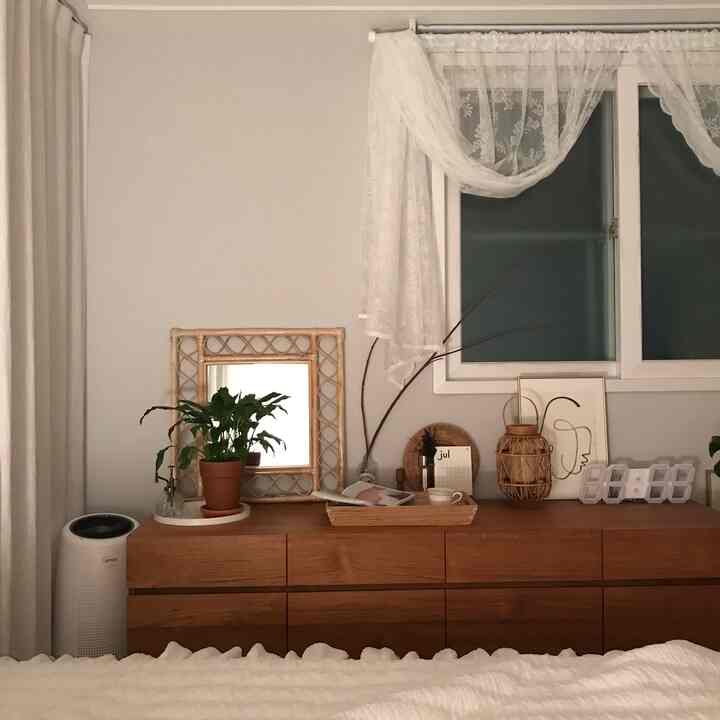 White and brown toned bedroom featuring teak wood sideboard, rattan mirror, and lace curtains creating a cozy nighttime atmosphere