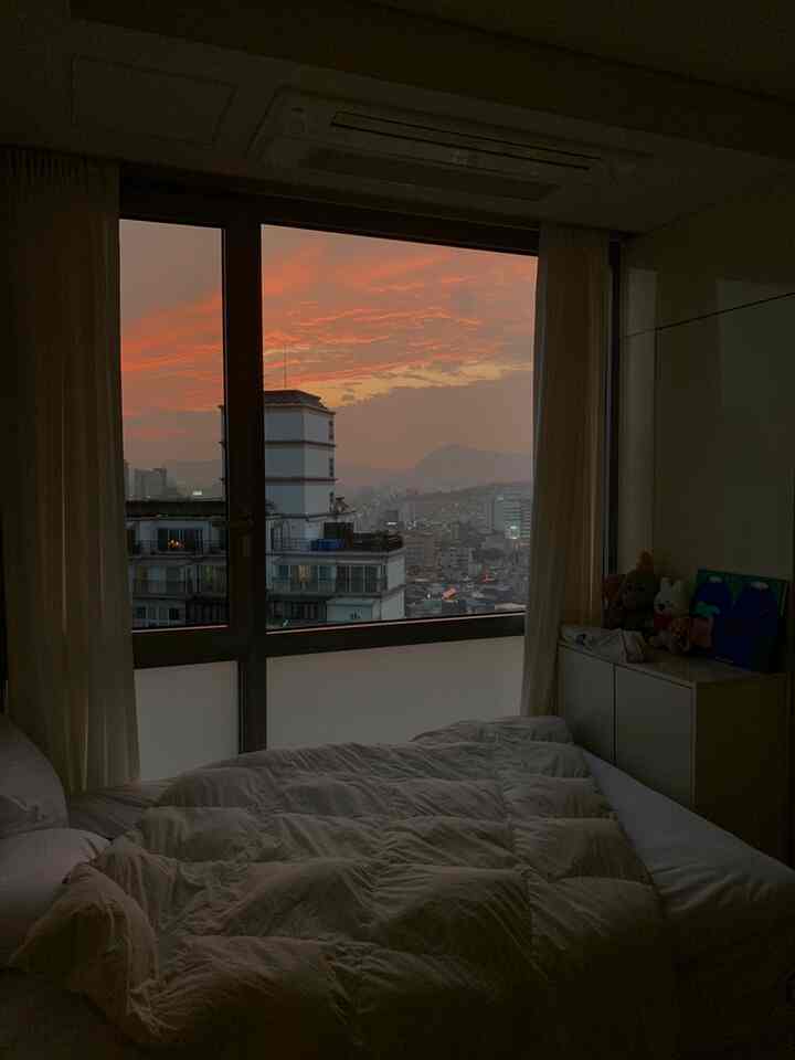 White-toned bedroom illuminated by sunset glow, featuring plush bedding and a cityscape view through the window