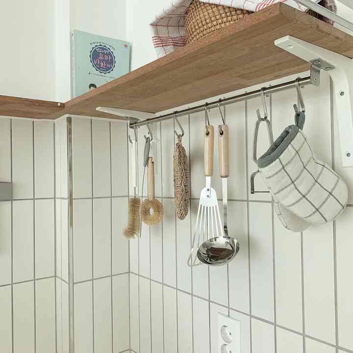 Natural-colored mulbau wood shelf and white tiled wall in kitchen area featuring hanging cooking utensils