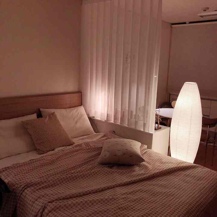 ベージュとホワイトのトーンのワンルーム寝室、中央にベッドとクッション、右側に明るいフロアランプが配置された温かみのあるミニマルなインテリア