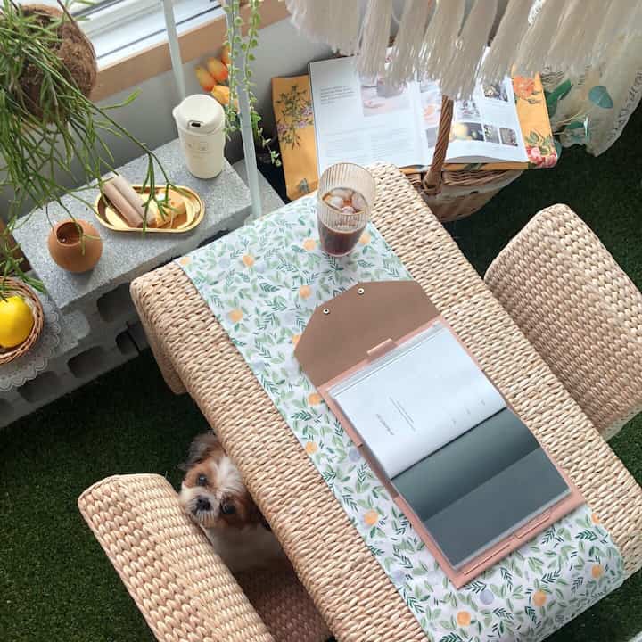 Natural-toned balcony space featuring rattan table, artificial grass floor, and a small dog creating a cozy home cafe vibe