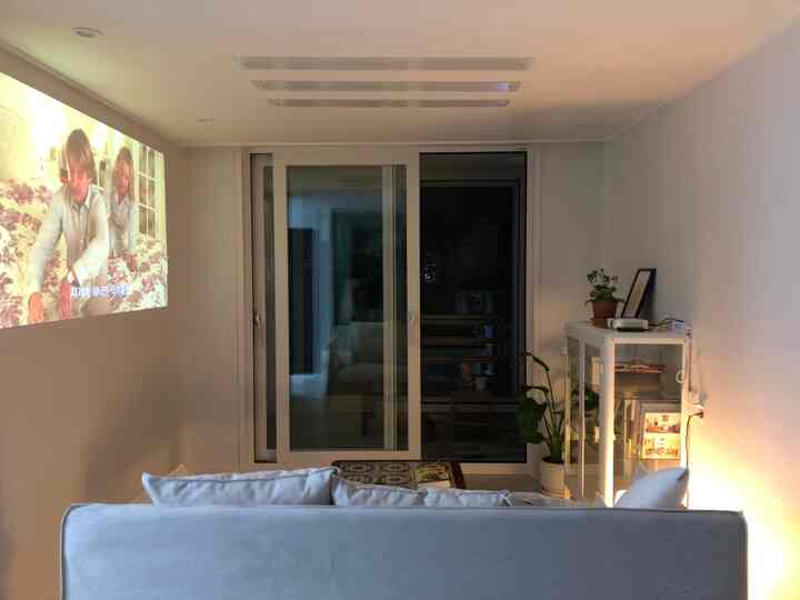 ホワイトトーンのリビング、壁にプロジェクター映像、クリーム色のソファ、白い収納家具と植物が配された温かみのある空間