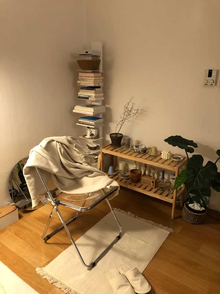 ベージュと木目調のナチュラルな一人暮らしの読書スペース、透明なアームチェアと木製の棚にキャンドルや植物が配置されたシンプルなインテリア