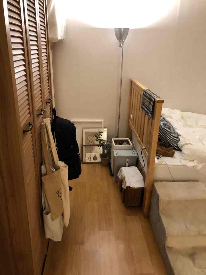 Natural-tone small bedroom featuring a wardrobe as room divider, wooden bed frame, and floor lamp creating a cozy atmosphere