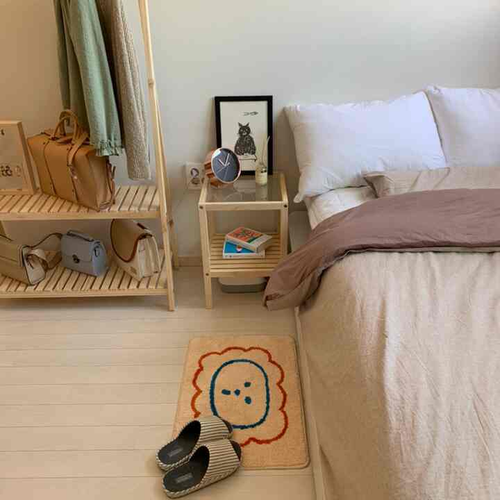 Natural color toned studio bedroom featuring a bed with bedding, wooden nightstand and open wooden shelf, cozy foot mat on floor