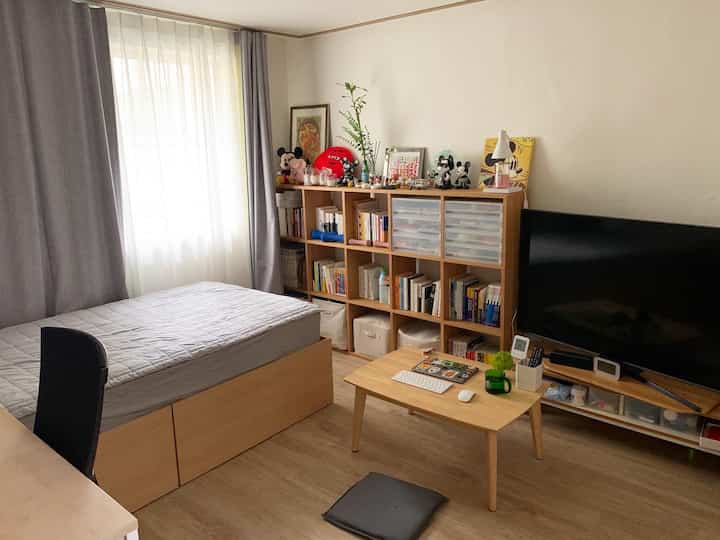Natural bedroom with white walls and wood-tone furniture for a single household, featuring storage bed, bookshelf, TV, and curtains