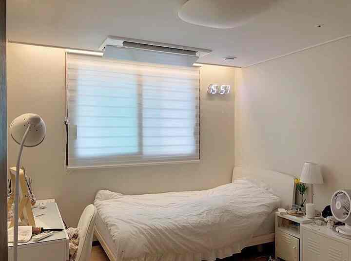 White and beige toned studio bedroom featuring a central bed, table lamp, and digital wall clock in a tidy personal space