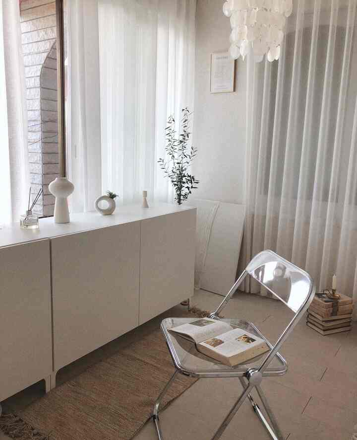 White and natural toned living room featuring a transparent chair, white cabinet, and sheer curtains with abundant natural light in a simple modern style