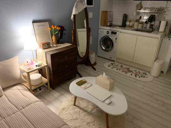 白と木目調が調和したワンルームのリビング、チェック柄ソファと楕円形のテーブル、テーブルランプと花瓶が配置された温かみのある空間