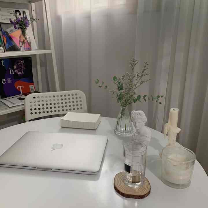 柔らかな白とベージュのトーンのホームオフィス、白いテーブル上にMacBook、石膏像、キャンドル、植物が配置されたシンプルなインテリア