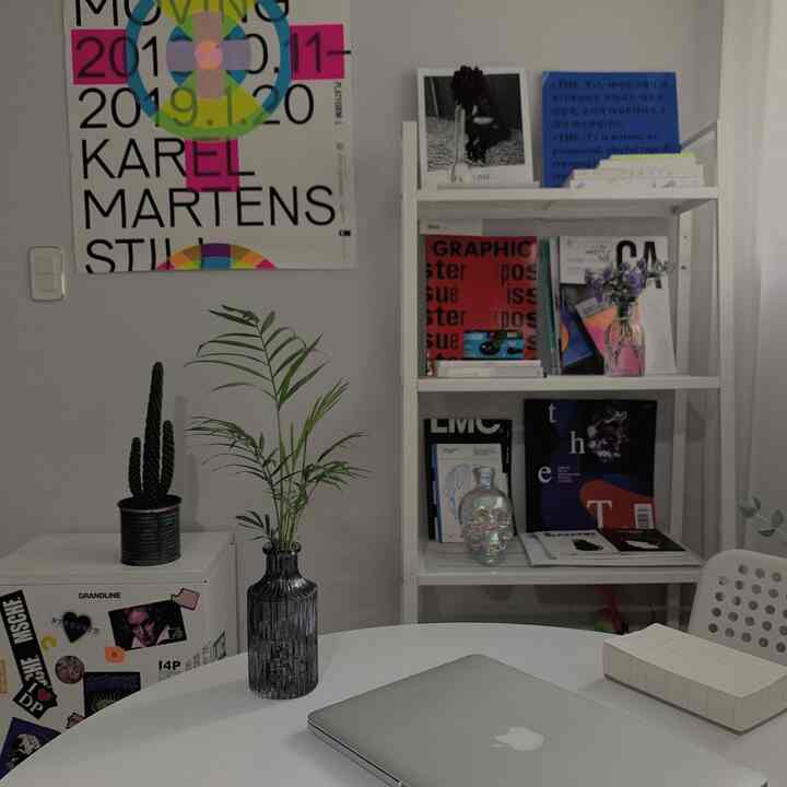 A white and gray toned studio apartment with a dining table featuring a laptop and plant, surrounded by bookshelf and posters, showcasing a simple home office.