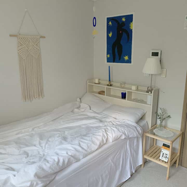 A white and natural tone bedroom featuring a bed, nightstand, macrame wall hanging, and fabric poster creating a cozy atmosphere