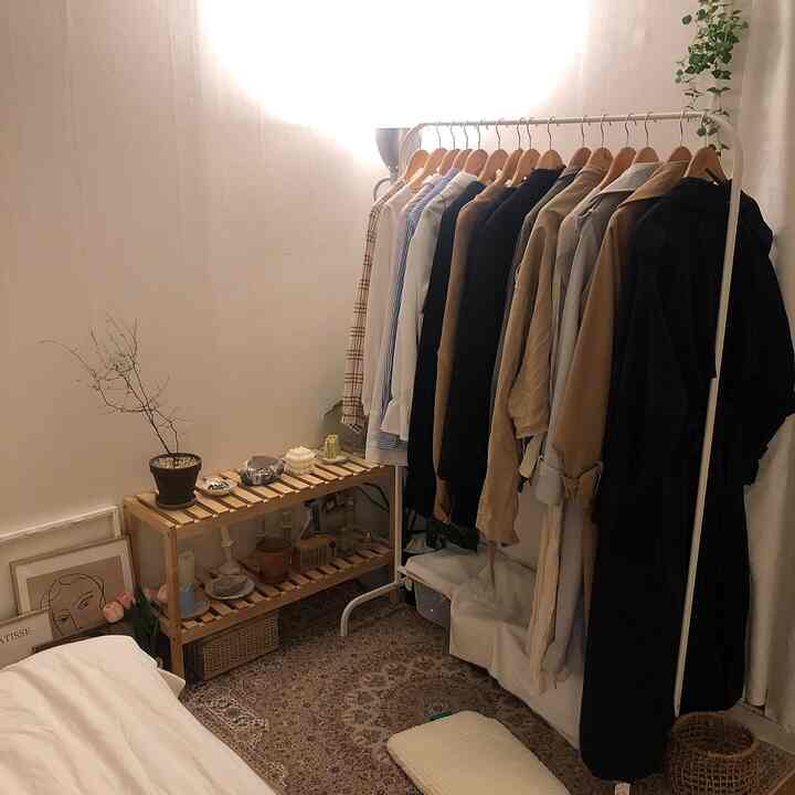 ナチュラルな色合いの一人暮らしの空間で、白いハンガーラックにさまざまな服が掛かり、木製の棚とペルシャラグが調和した雰囲気のあるインテリア