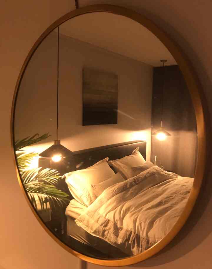 Round mirror reflecting a modern bedroom with black bed, white bedding, and warm pendant lighting