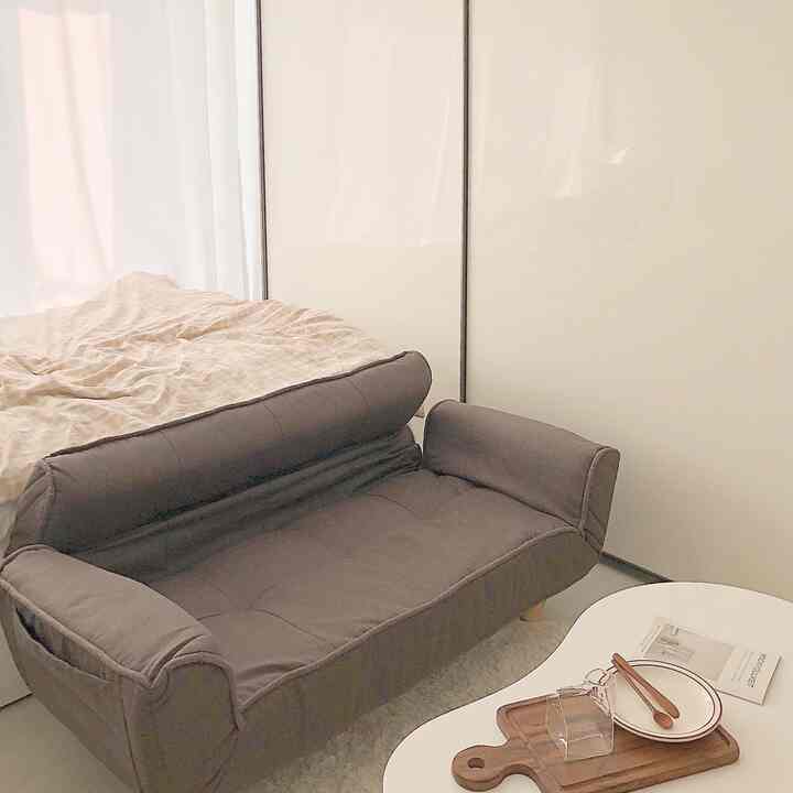 Beige-toned studio bedroom featuring a foldable gray sofa bed, round rug, and white wooden table, with a simple and cozy atmosphere