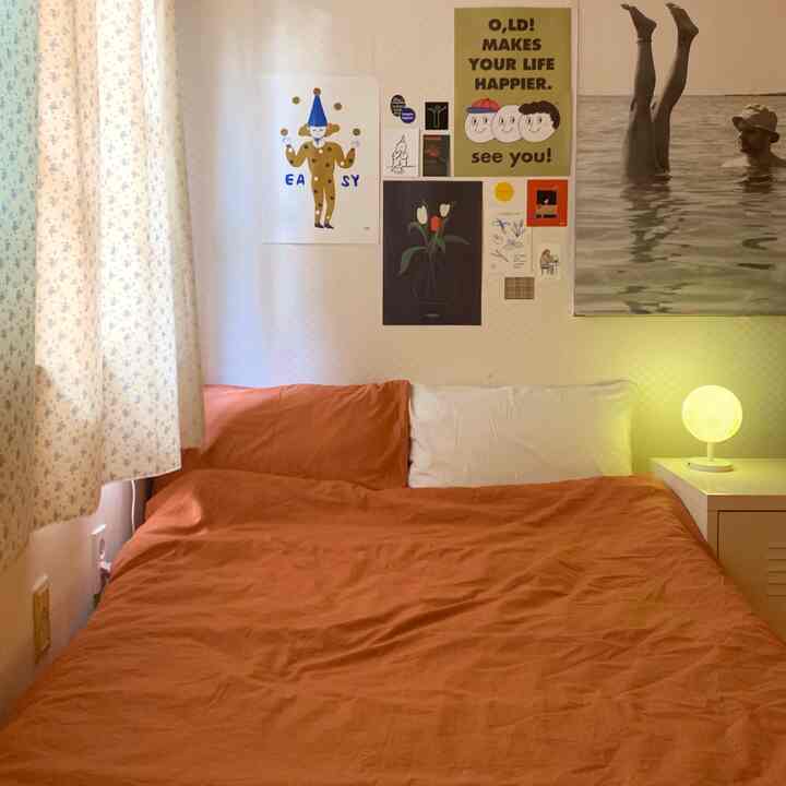 暖かいオレンジ色の寝具と多様なポスターが配置されたワンルーム寝室、ムードランプが居心地の良い雰囲気を演出する空間