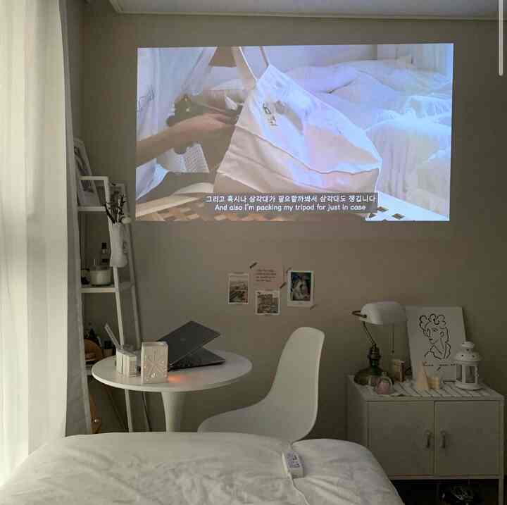White-toned small bedroom featuring a round table with laptop, shelves, and a projector screen creating a cozy home office space
