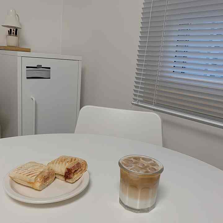 Small white-toned dining room featuring a white table and chair, gray blinds, and a neat home cafe vibe with iced latte and sandwiches