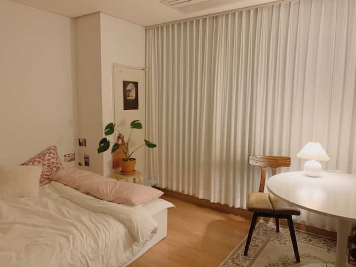 Cozy bedroom with white curtains and wooden floor featuring a bed, wooden chair, and table lamp creating warm atmosphere