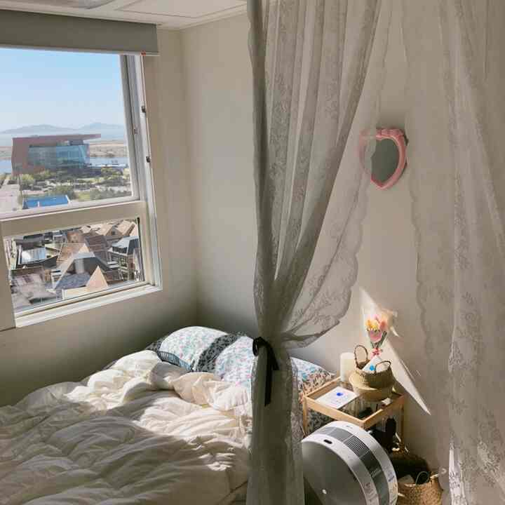 ホワイトトーンの小さな寝室、柔らかなレースカーテンと木製のナイトテーブルが温かみのある雰囲気を醸し出す空間