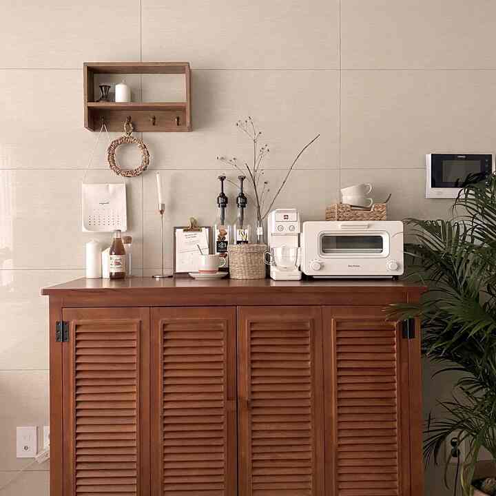 ナチュラルな木目調とベージュの壁のあるおうちカフェ、コーヒーマシンやトースターオーブンが置かれたルーバーキャビネットと植物が配置された雰囲気のあるインテリア