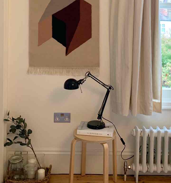 Natural-toned small room corner with wood stool, black table lamp, beige curtains, and geometric wall decor featuring white walls