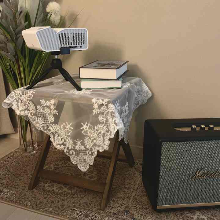 ブラウンとホワイトのトーンの小さな空間、レースのテーブルクロスをかけた木製のナイトテーブルにプロジェクターと本が配置された雰囲気のあるインテリア