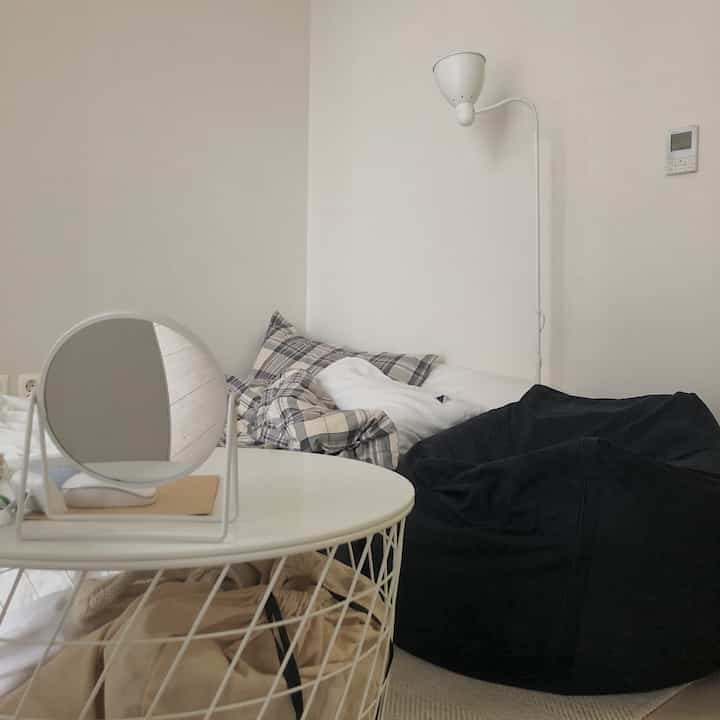 A simple white-toned small room featuring a bed, bean bag sofa, round table, and floor lamp creating a cozy atmosphere