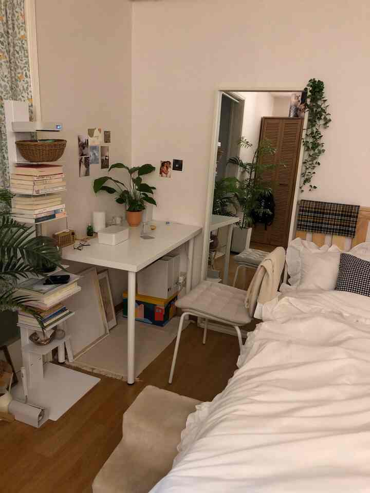 ホワイトとブラウンの色調の寝室、ベッド横に置かれたデスクと全身鏡、観葉植物が配置された落ち着いた空間