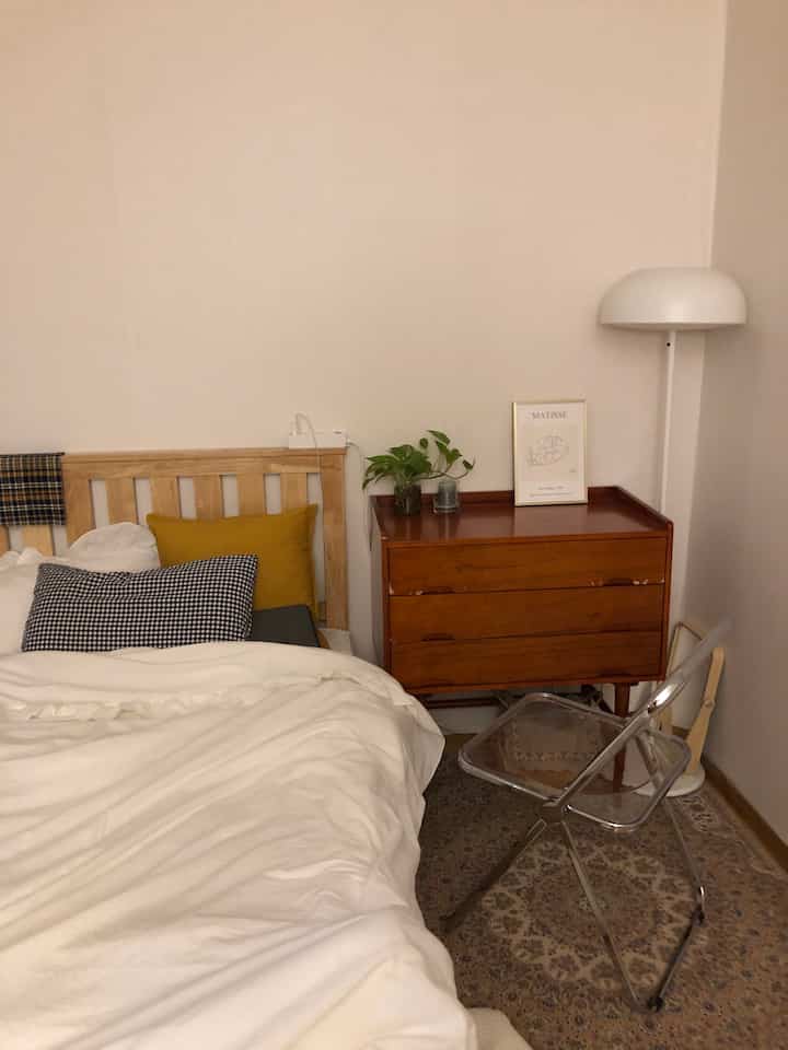 白を基調とした寝具と木目調家具が配置されたシンプルで自然な雰囲気の寝室、透明な椅子とフロアランプ付き