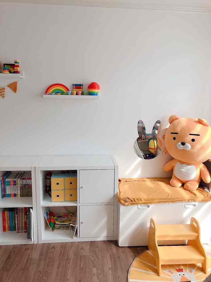 Bright and cozy kids' room with white walls and wood-tone flooring, featuring white bookshelves, a yellow two-step stool, rabbit-shaped safety mirror, and a character plush toy