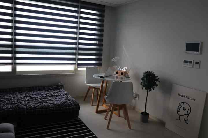 白を基調とした小さな寝室、黒のストライプカーテン、丸テーブルと椅子、ベッドと額縁が配置されたシンプルなインテリア