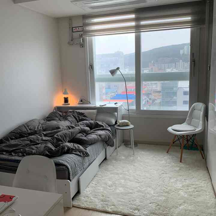 White and gray toned studio apartment bedroom featuring simple bed, lighting, small nightstand, and chair in a cozy space
