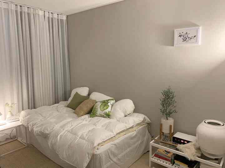 A small bedroom with beige walls and white bedding featuring green cushions and a plant, creating a natural and cozy atmosphere