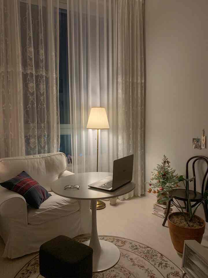 Soft white-toned studio living room featuring a white sofa, round table, warm floor lamp, and decorated Christmas mini tree creating a cozy atmosphere