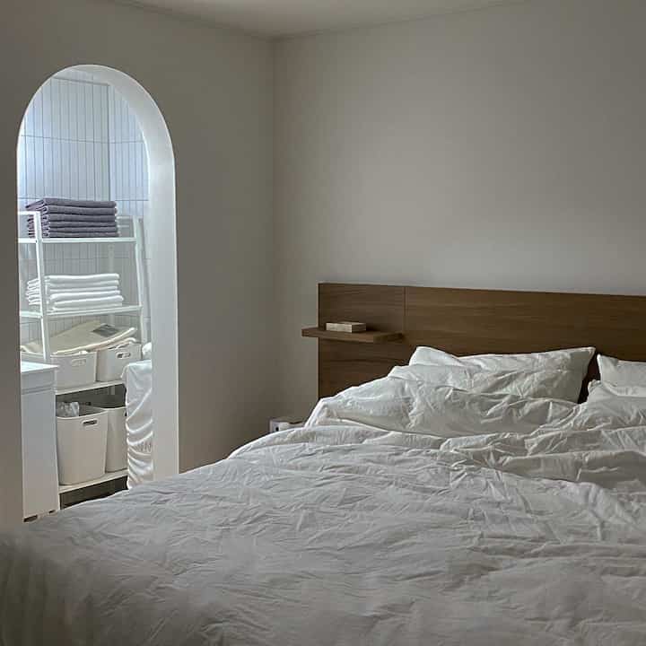 白と木目調の寝室、アーチ型の入り口からタオルと棚が配置されたホテルライクなバスルームが見える空間