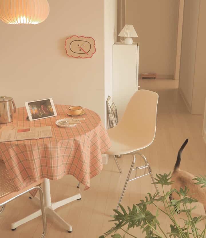 暖かいナチュラルトーンのキッチン兼ダイニングルーム、円形のダイニングテーブルと白いチェア、壁掛け時計、猫がいる雰囲気のある空間