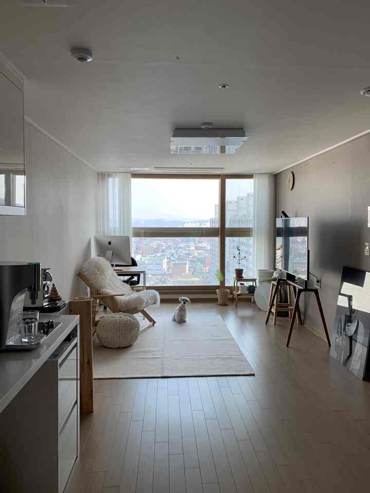 White and wood tone living room with desk by the window, armchair, knit stool, and a small dog on the center rug, simple atmosphere